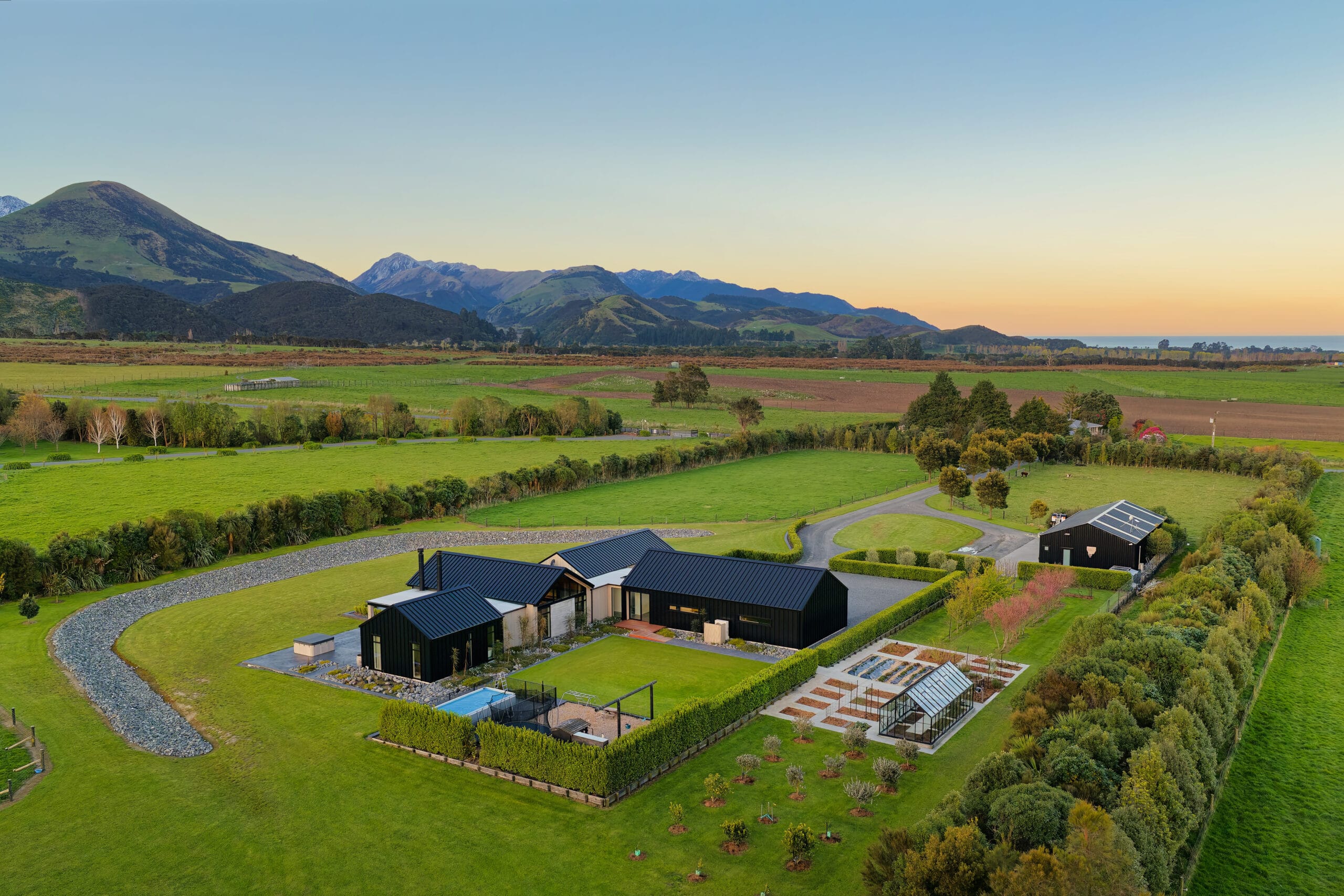 Aerial view of green farmland with modern dark-roofed houses, hills, and sunset sky.