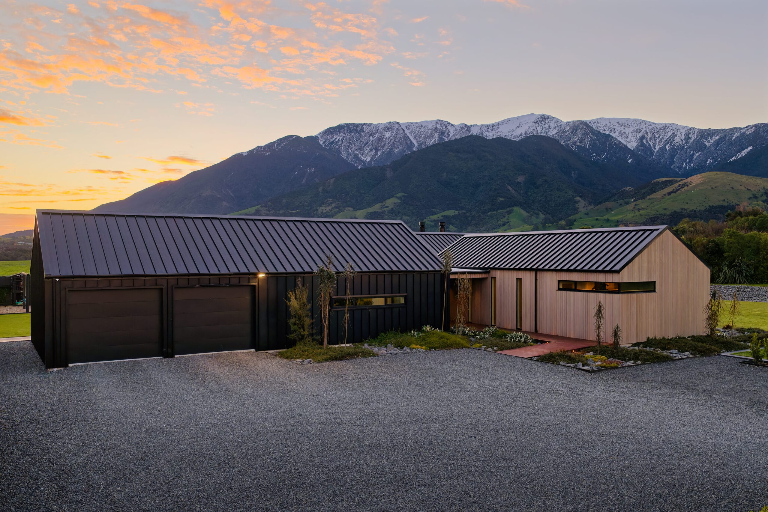 Modern building with dark metal roof and light wood siding, set in gravel lot with mountains in the distance.