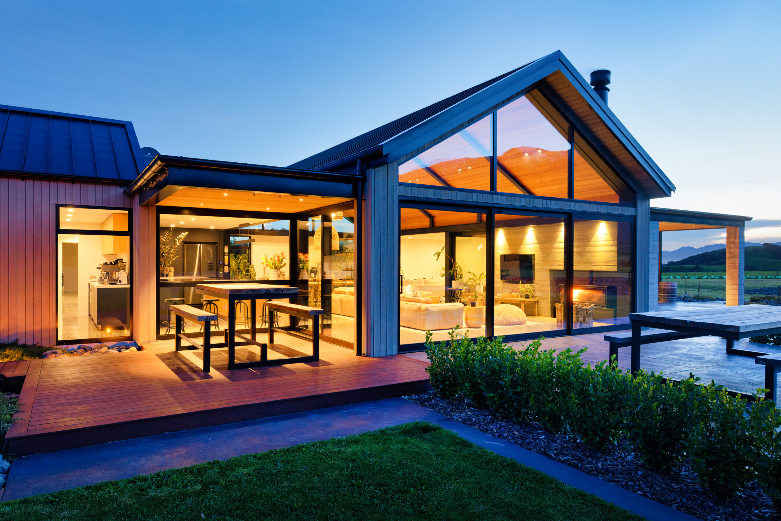 Modern glass-walled house with warm interiors visible at dusk, wooden deck.