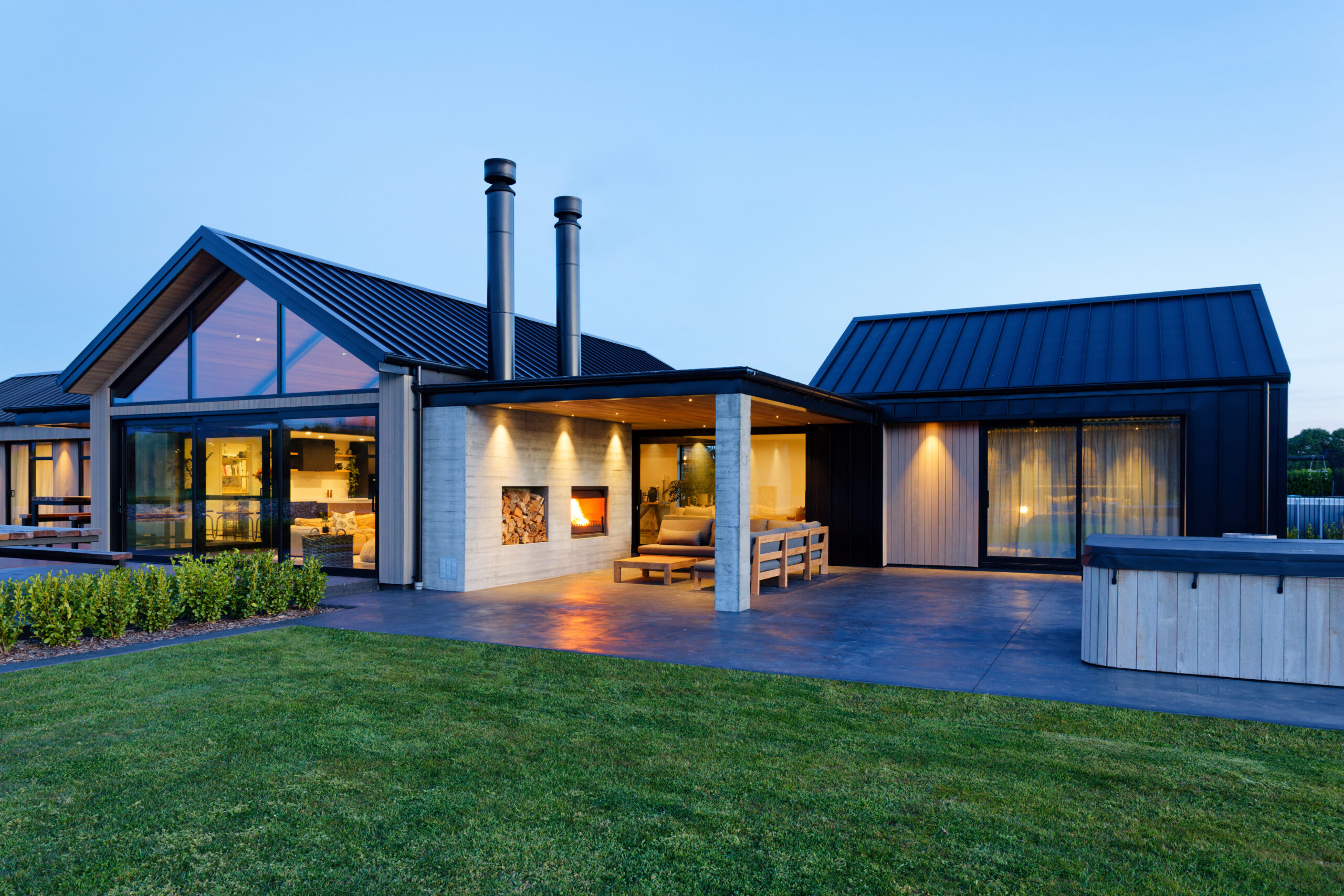Modern house with dark metal roof, large glass walls, and outdoor seating at dusk.
