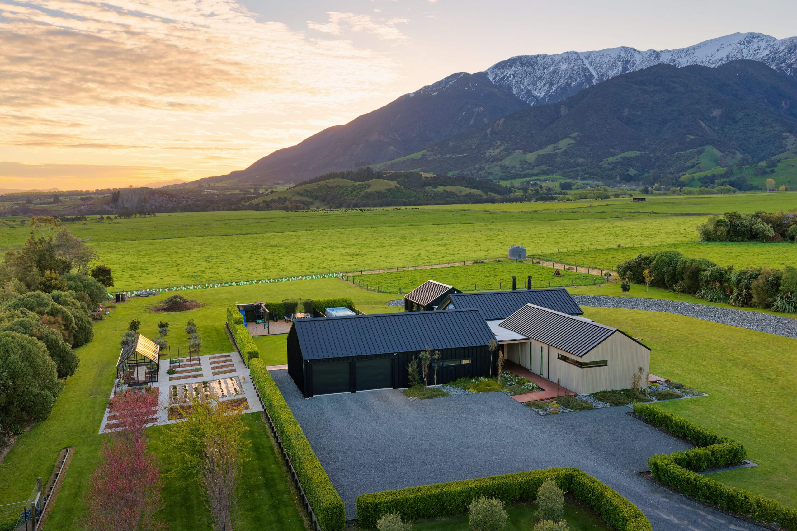 Modern house with dark roof, light walls, sprawling green yard, and snow-capped mountains at sunset.