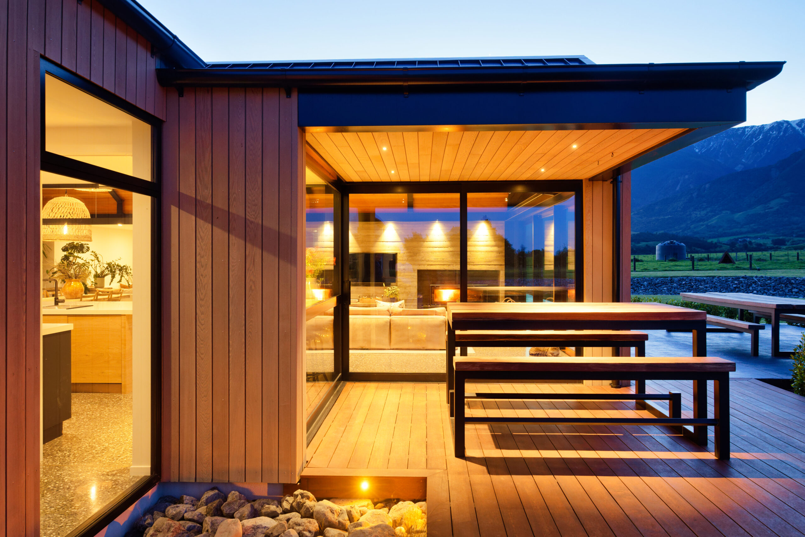 Modern house with wood siding, glass doors, warm interior, and mountain landscape view.