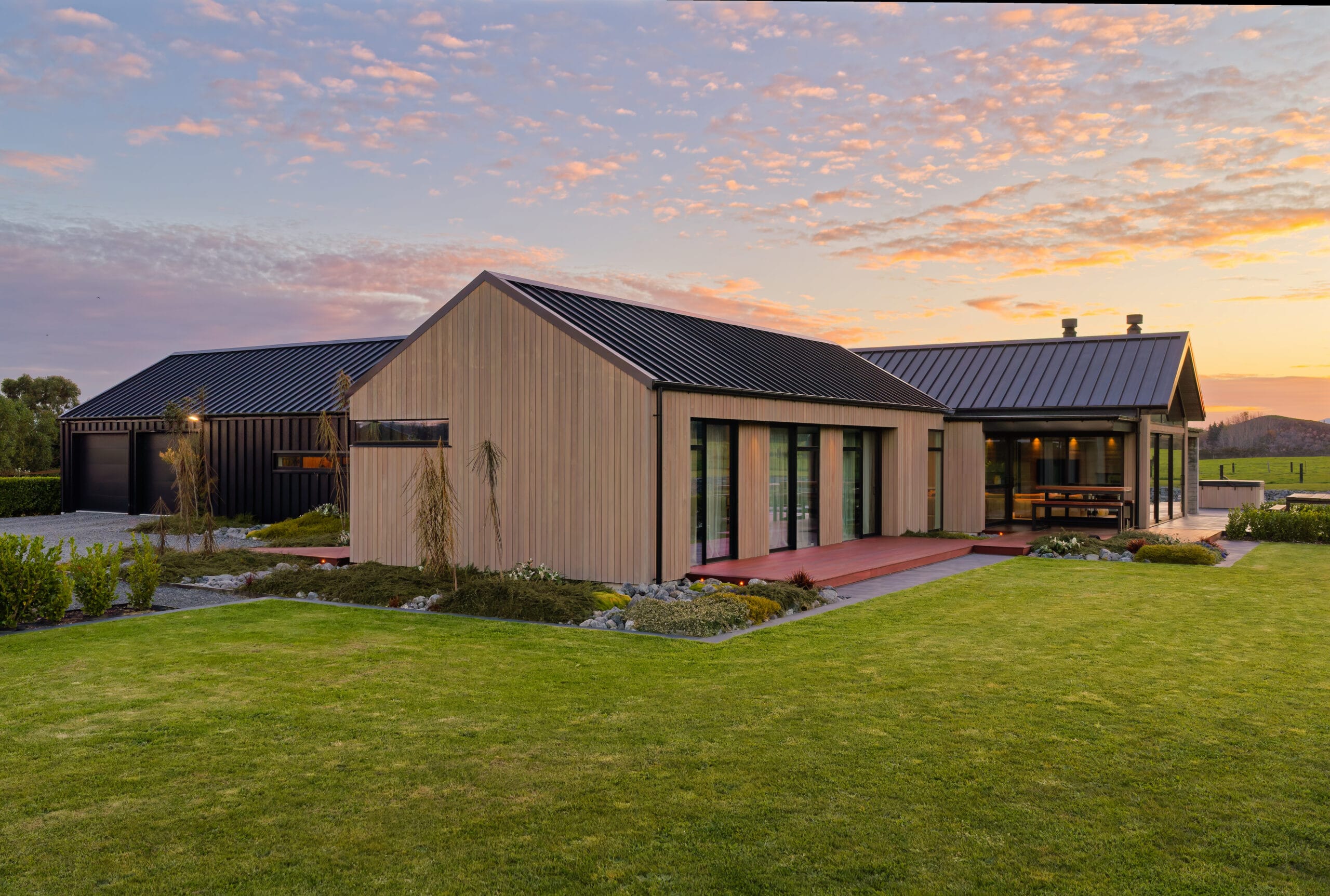 Modern single-story house with metal roof, light siding, deck, and large glass doors at sunset.