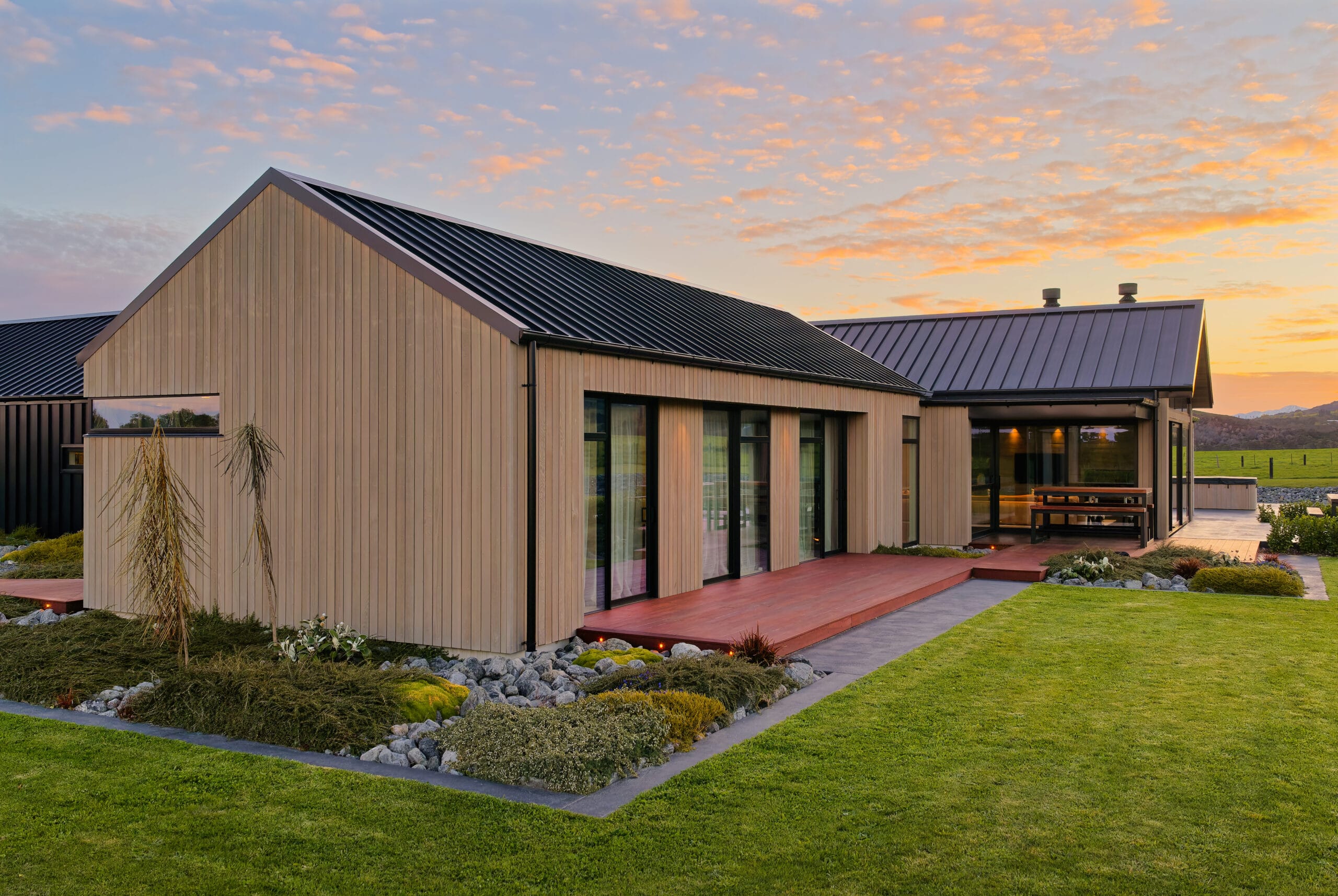 Modern single-story house with wood siding, black metal roof, glass doors, and patio at sunset.