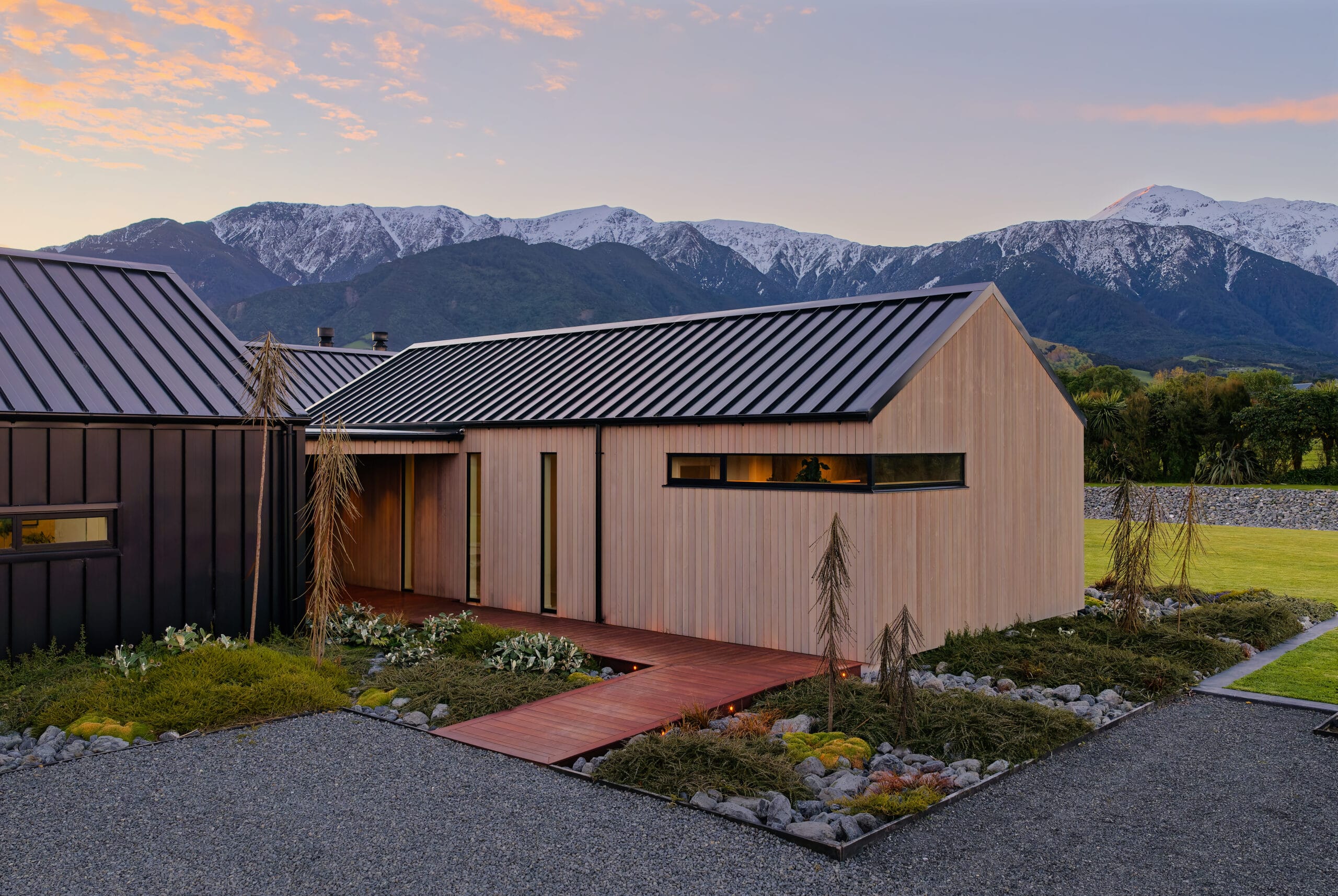 Modern wooden houses with metal roofs against snow-capped mountains at dusk.