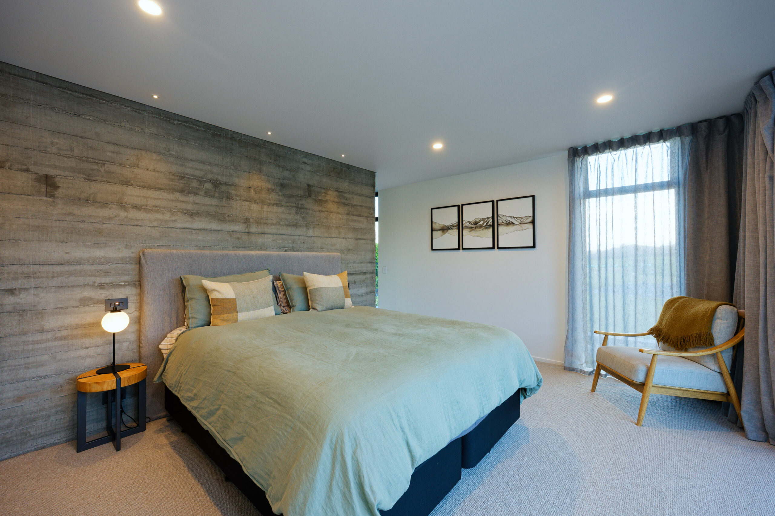 Bedroom with grey wood accent wall, teal bedding, and three-panel mountain art; large window with sheer curtains.