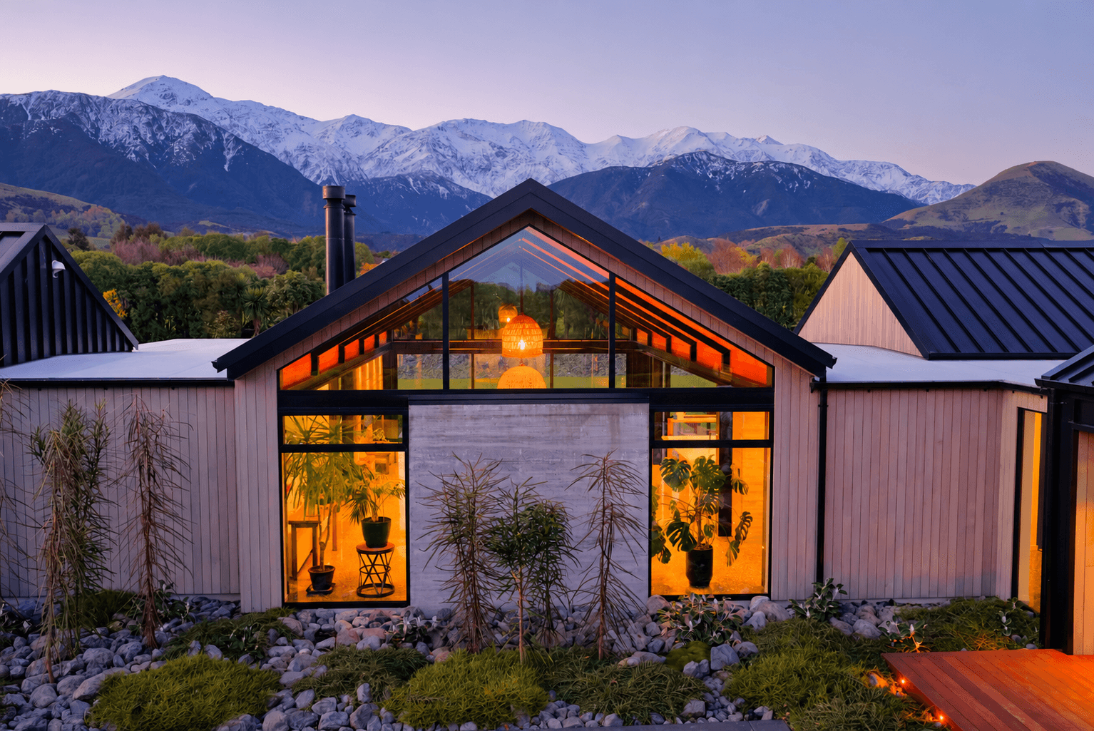 Modern glass-walled house with triangular roof, warm interior lights, rocky garden, and snow-capped mountains in the distance.
