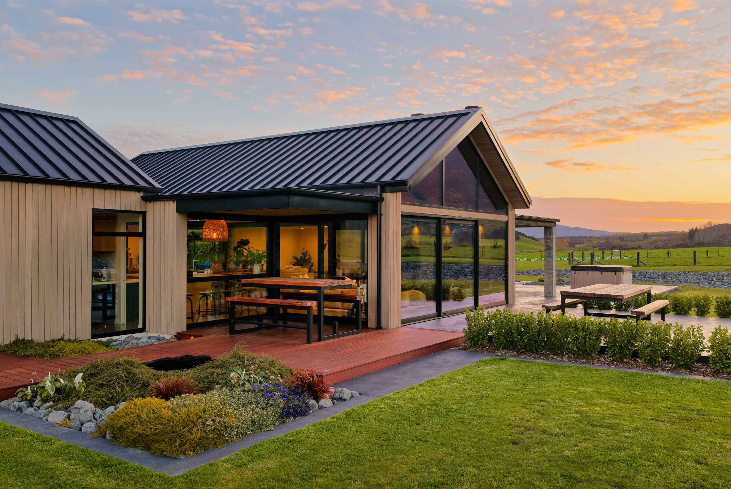 Modern house with metal roof and large glass windows, sunset-lit yard, wooden deck, and picnic tables.
